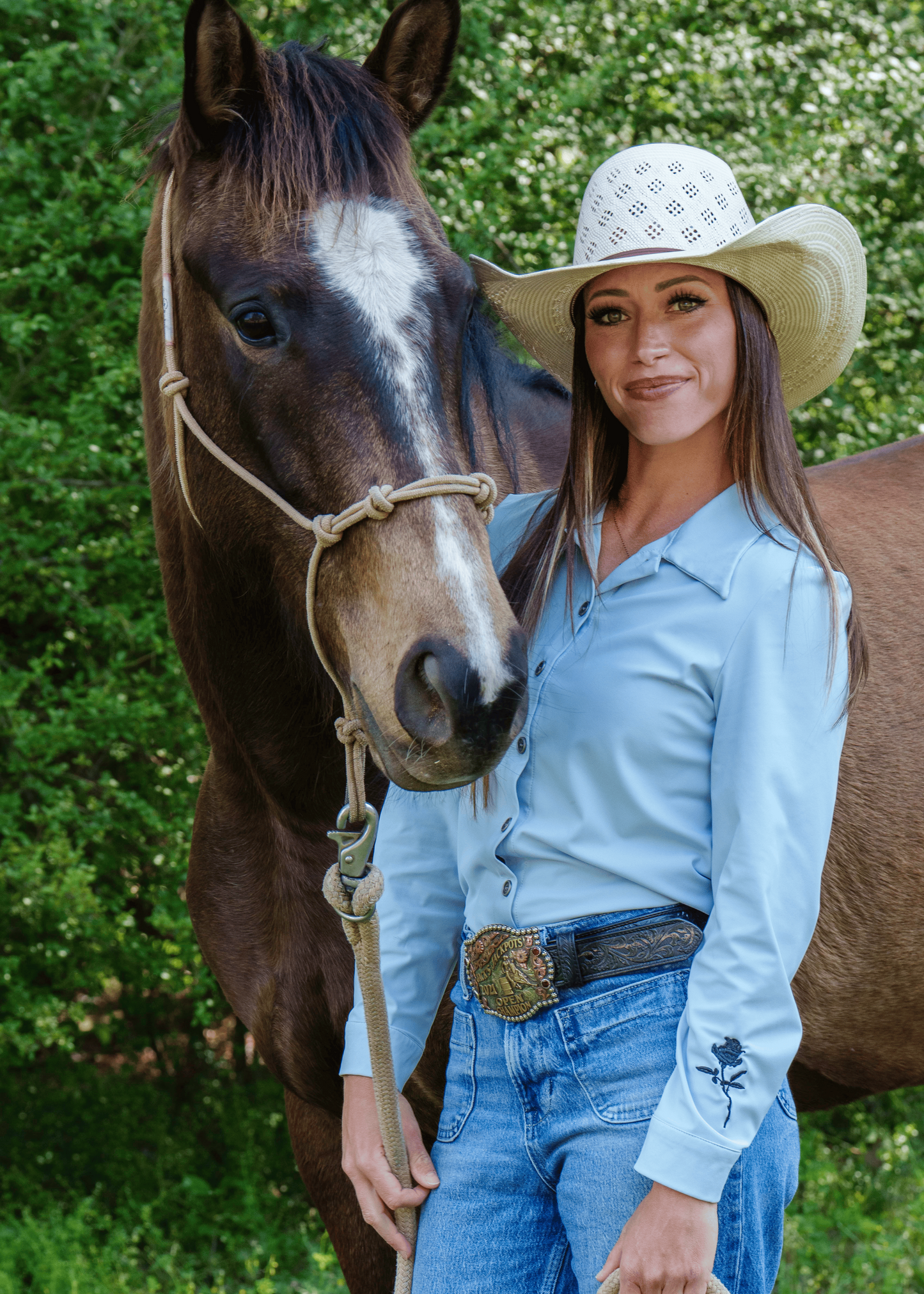 Woman in a cowboy hat and blue shirt standing next to a horse outdoors.