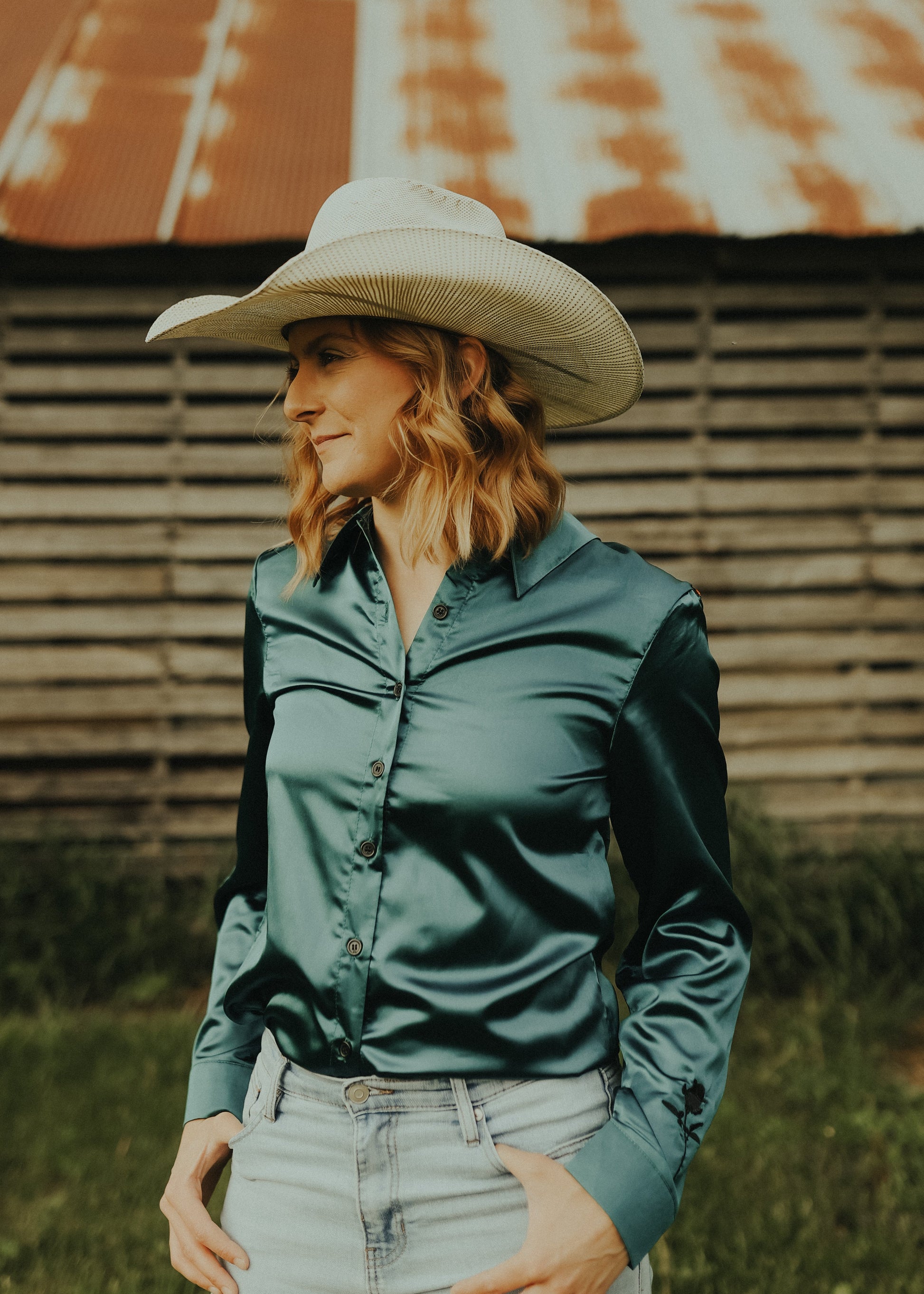Woman wearing a cowboy hat and green shirt standing in front of a rustic wooden building.